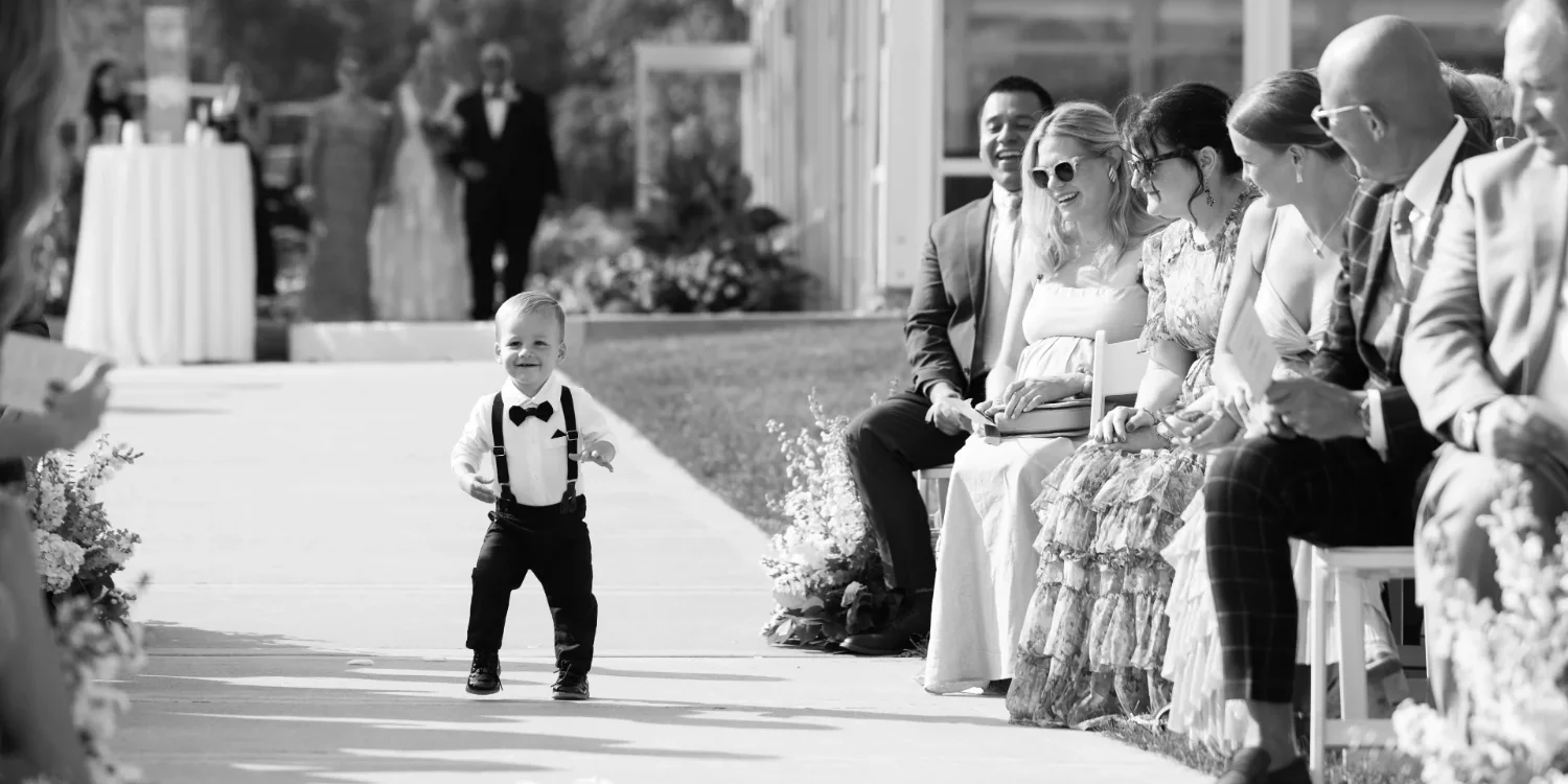 A happy baby in suspenders being admired by sitting wedding goers. Photo shot by C.Mae Photography.